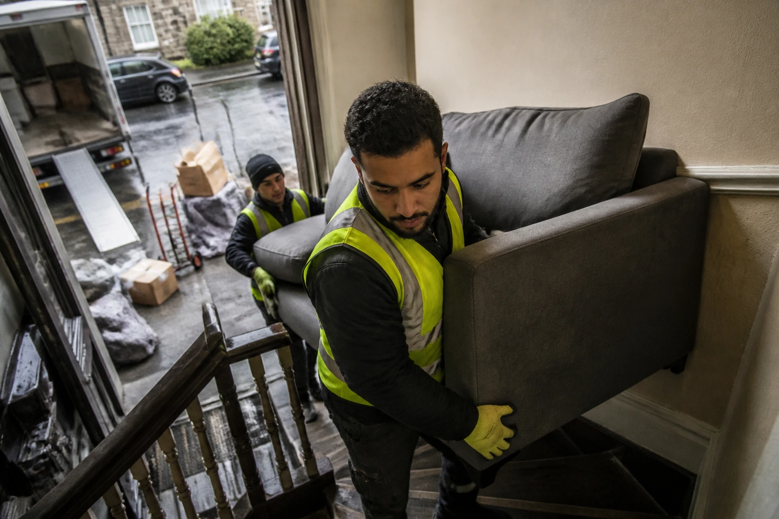 Mover guiding a sofa through a narrow stairwell on a rainy day.